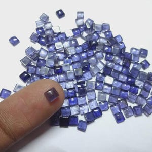 Peut inclure: Un ensemble de petits gemmes carrées bleues, dispersées sur une surface blanche. Une gemme repose sur un doigt. Les gemmes varient en nuances de bleu, du clair au foncé, et semblent translucides.