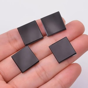 May include: Four square, black onyx gemstones are shown. Each stone is a polished, solid black colour and has a smooth, reflective surface. The gemstones are held in a hand, showcasing their size and shape. These stones could be used for jewellery making or other crafts.