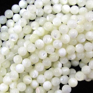 May include: A close-up shot of a pile of luminous, round, off-white beads. The beads have a pearlescent sheen, reflecting light. The beads are uniform in size and shape, suggesting they are for jewelry making or crafts.
