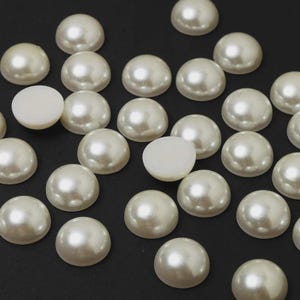 May include: A collection of ivory-colored, half-round faux pearls scattered on a dark surface. The pearls have a lustrous, pearlescent sheen, with a flat, unpolished back. These craft supplies are ideal for jewelry making and embellishments.