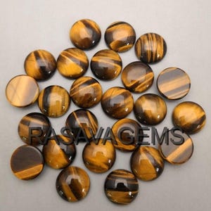 Peut inclure: Une collection de pierres précieuses œil de tigre polies et rondes. Les pierres présentent un mélange de bandes brun doré et brun foncé, avec une finition brillante. Les pierres sont disposées sur une surface gris clair.