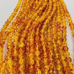 May include: Close-up of multiple strands of amber-coloured beads. The beads are round and have a speckled, translucent appearance with red and orange inclusions. The beads are strung together, creating a textured and layered effect.