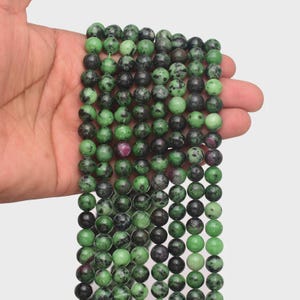 Puede incluir: Hilo de cuentas redondas de piedras preciosas en tonos verdes y negros, con inclusiones rojas ocasionales. Las cuentas se sostienen en una mano, mostrando su superficie pulida y variaciones naturales. Las cuentas tienen aproximadamente 8 mm de diámetro.