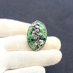 Puede incluir: Un anillo ovalado con una piedra verde de motas negras, acentuado por un aplique de metal plateado con forma de enredadera con pequeñas bayas y hojas. El engaste está pulido y sigue el contorno ovalado de la piedra central.