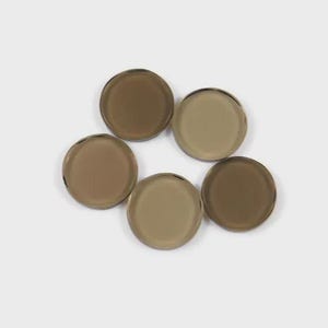 Könnte beinhalten: Sechs runde Glasscheiben in verschiedenen Braun- und Beigetönen liegen auf einer weißen Oberfläche. Die Glasscheiben haben eine reflektierende Qualität, und die Farben reichen von hellem Beige bis zu einem dunkleren, kaffeeähnlichen Farbton.