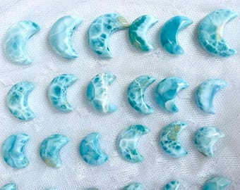 Puede incluir: Una colección de piedras preciosas pulidas de larimar en forma de media luna. Las piedras varían en tonos de azul y blanco, con patrones únicos. Las lunas están dispuestas sobre una superficie blanca texturizada, mostrando su belleza natural.