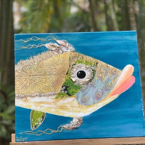 Puede incluir: Una pintura colorida y mixta de un pez con un cuerpo azul, verde y dorado. El pez tiene un ojo grande y redondo y una boca rosa. El fondo es de un azul vibrante.