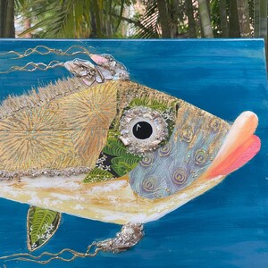 Puede incluir: Una pintura abstracta de un pez colorido sobre un fondo azul. El pez est&aacute; hecho de varios materiales, incluyendo madera, tela y purpurina. El ojo del pez es un c&iacute;rculo grande negro con un anillo blanco alrededor.
