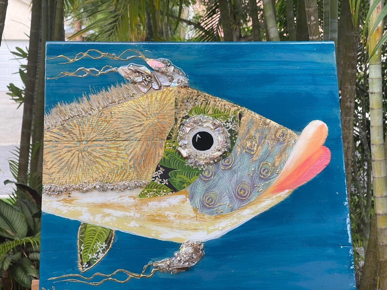 Puede incluir: Una pintura abstracta de un pez colorido sobre un fondo azul. El pez est&aacute; hecho de varios materiales, incluyendo madera, tela y purpurina. El ojo del pez es un c&iacute;rculo grande negro con un anillo blanco alrededor.