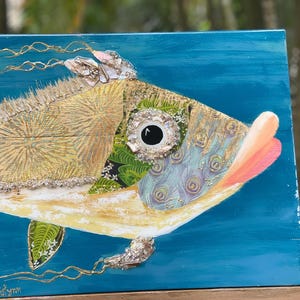 Puede incluir: Una pintura colorida y mixta de un pez con un ojo grande e iridiscente. El pez est&aacute; pintado sobre un fondo azul y tiene una boca rosa. El pez est&aacute; hecho de varios materiales, incluyendo tela, papel y pintura.