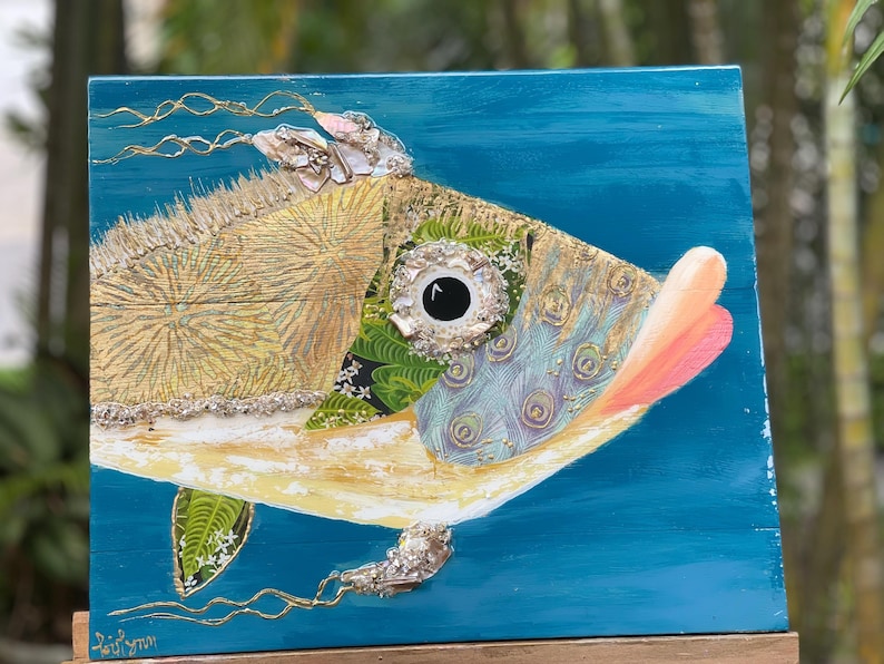 Puede incluir: Una pintura colorida y mixta de un pez con un ojo grande e iridiscente. El pez est&aacute; pintado sobre un fondo azul y tiene una boca rosa. El pez est&aacute; hecho de varios materiales, incluyendo tela, papel y pintura.
