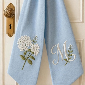 May include: A light blue hand towel featuring white embroidered hydrangea flowers on the left side and a large white embroidered script letter M accompanied by a small green and white floral sprig on the right side of the fabric.