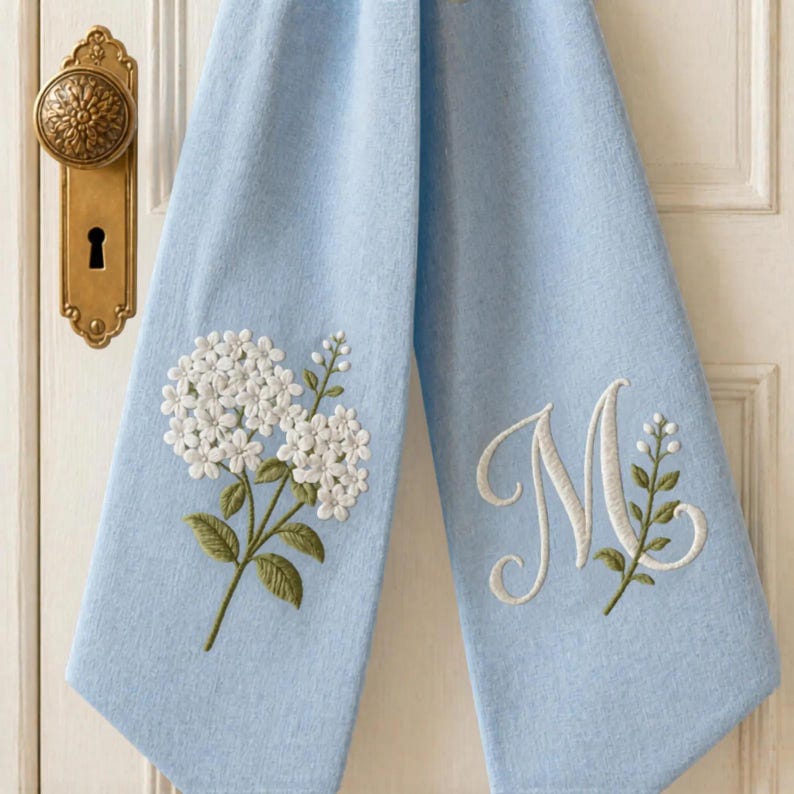 May include: A light blue hand towel featuring white embroidered hydrangea flowers on the left side and a large white embroidered script letter M accompanied by a small green and white floral sprig on the right side of the fabric.