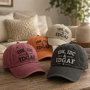 May include: Three baseball caps in maroon, orange, and charcoal gray, plus one beige cap in the back. Each cap features embroidered black or white text that reads IDK, IDC & IDGAF with horizontal lines and a small star symbol centered below the text.