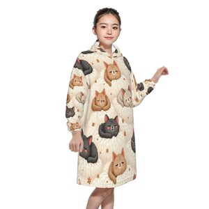 Young woman wearing a cozy hoodie dress adorned with cute, sleeping cat illustrations in various colors on a soft, cream background. She looks content.