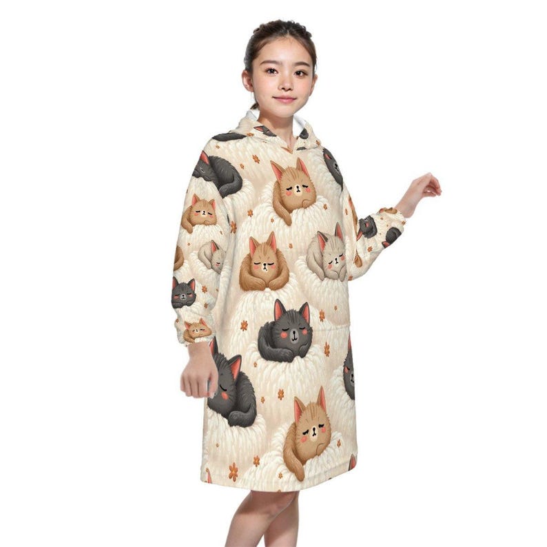 Young woman wearing a cozy hoodie dress adorned with cute, sleeping cat illustrations in various colors on a soft, cream background. She looks content.