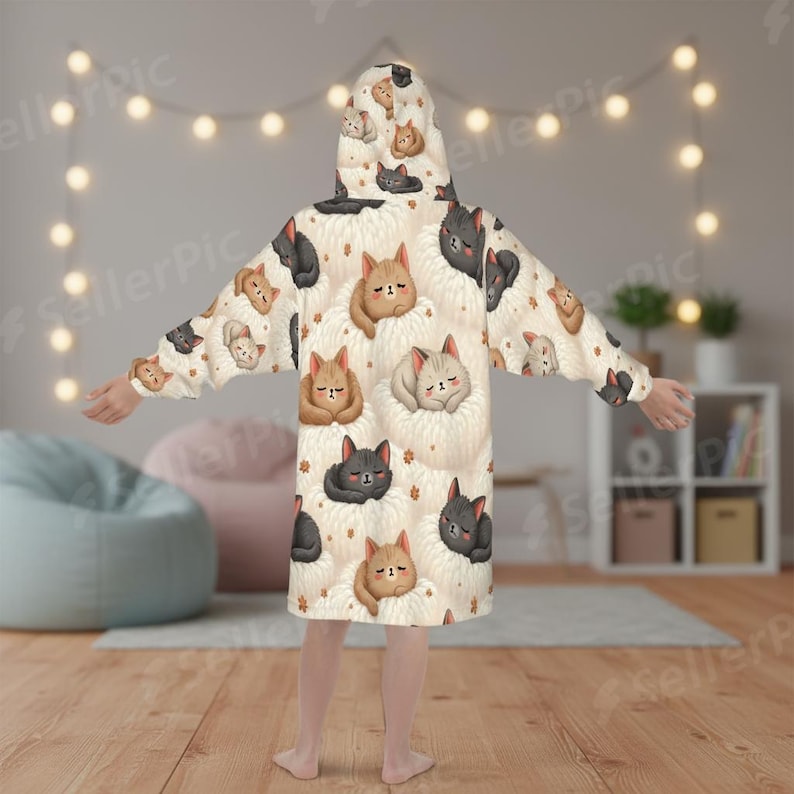 A person wearing a cozy, hooded blanket adorned with cute cat illustrations stands in a softly-lit room. String lights and beanbags enhance the cozy atmosphere.