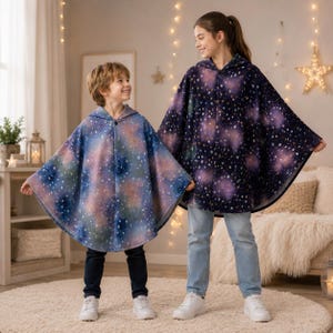 May include: Two hooded ponchos featuring a celestial galaxy print with stars, nebulae, and cosmic clouds. One poncho displays a lighter blue and pink gradient, while the other features a darker navy blue and purple base with a dense star pattern.
