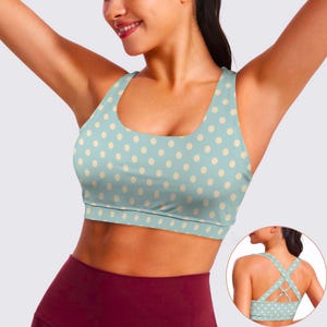 May include: A light blue sports bra with a cream-colored polka dot pattern. The bra has a scoop neck and a criss-cross back design. The model is wearing maroon leggings.