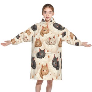 Young person wearing an oversized hoodie featuring cute, cartoon-style cats in various colors on a fluffy background, conveying a cozy, playful vibe.