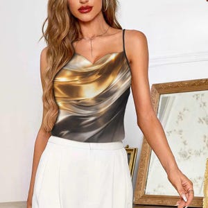 May include: Satin camisole top with a draped cowl neckline. The top features a metallic gold, silver, and black abstract design. The top has thin black spaghetti straps and is paired with white pants.