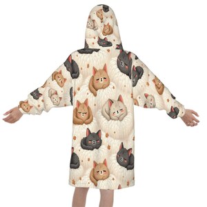 A person wearing a cozy hooded robe with a playful pattern of cartoon cats in brown, gray, and beige shades, set against a cream background. The cats appear fluffy and content, adding a warm, whimsical tone.