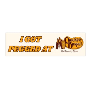 Peut inclure: Autocollant rectangulaire avec le texte "I GOT PEGGD AT" en lettres orange. Le logo Cracker Barrel est à droite, représentant un homme, un tonneau et les mots "Cracker Barrel Old Country Store."