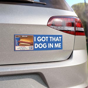 Könnte beinhalten: Ein blauer Aufkleber auf einem silbernen Auto mit dem Text "I GOT THAT DOG IN ME". Der Aufkleber zeigt auch ein Bild eines Hotdogs und einer Pepsi, mit dem Text "KIRKLAND" und "ALL BEEF HOT DOG UND 567 ml SODA."
