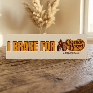 Peut inclure: Panneau rectangulaire avec le texte "I Brake For" en lettres orange. Le panneau présente un logo pour Cracker Barrel Old Country Store, avec une silhouette assise et un tonneau. Le panneau est sur une surface en bois.