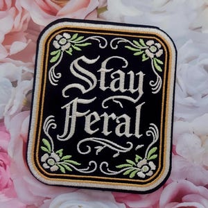 May include: A rectangular embroidered patch with a black background and a gold border. The text "Stay Feral" is stitched in white in a decorative font. The design features white and light green floral accents in the corners with scrolling lines.