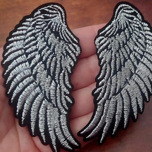 Puede incluir: Un par de parches bordados con forma de alas de ángel, con detalles de hilo plateado metálico sobre una base de tela negra sólida. El diseño muestra intrincados patrones de plumas con un contorno definido, creando un aspecto de alto contraste.
