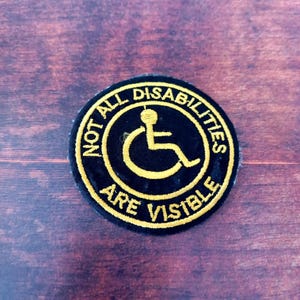 Peut inclure: Écusson rond noir avec texte brodé jaune et symbole de fauteuil roulant. Le texte dit "NOT ALL DISABILITIES ARE VISIBLE". L'écusson est sur une surface en bois foncé.