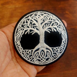 May include: A circular black patch with a white embroidered Tree of Life design. The tree has detailed roots, trunk, and branches. The patch is held in a hand, highlighting the intricate embroidery.