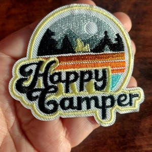 Op de afbeelding: Geborduurde patch met een campingthema. De patch toont een berglandschap met een volle maan, bomen en de tekst "Happy Camper" in een gestileerd lettertype. De patch heeft een gele rand en een kleurenpalet van zwart, oranje, turquoise en grijs.