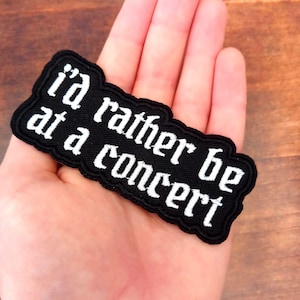 May include: A black embroidered patch with the white text "I'd rather be at a concert". The patch has a black border and a rectangular shape with rounded corners. The patch is held in a hand.