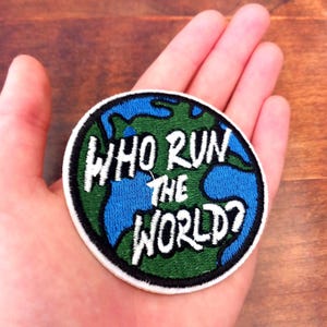 May include: Embroidered patch featuring a stylized globe with blue oceans and green continents. The patch has the text "WHO RUN THE WORLD?" in white, outlined in black, on a white background.