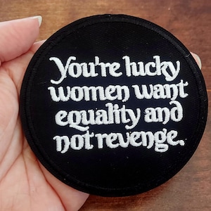 May include: A circular black patch with white embroidered lettering. The text says "You're lucky women want equality and not revenge." The patch has a stitched edge and is designed to be attached to fabric.