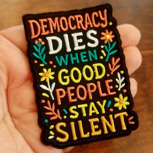 Pode incluir: Patch bordado preto com as palavras "DEMOCRACY DIES WHEN GOOD PEOPLE STAY SILENT" em laranja, branco, verde-azulado e amarelo. O patch é decorado com desenhos florais e de folhas.