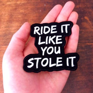 Pode incluir: Patch bordado preto com as palavras "RIDE IT LIKE YOU STOLE IT" em letras brancas. O patch é segurado em uma mão contra um fundo de madeira.
