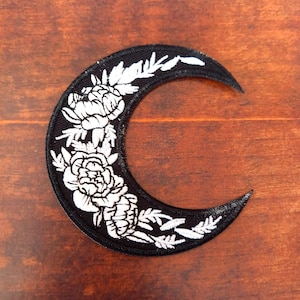 May include: Black crescent moon patch with white floral embroidery. The patch features detailed white flowers and leaves against a black background. The patch is likely for clothing or accessories.