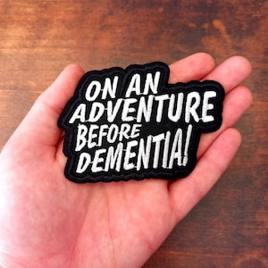 May include: A black embroidered patch with white text: "ON AN ADVENTURE BEFORE DEMENTIA!". The patch is held in a hand against a brown wooden background. The patch has a black border with white stitching.