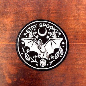 May include: A round, black patch with white embroidered details. The patch features a bat with outstretched wings, a crescent moon, stars, and the words "STAY SPOOKY" in a circular design. The patch is likely for clothing or accessories.