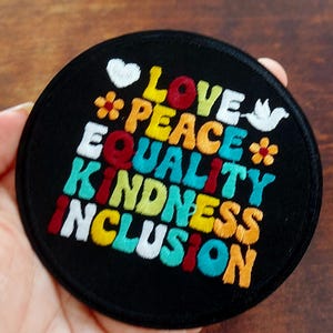 May include: A circular black embroidered patch featuring the words LOVE, PEACE, EQUALITY, KINDNESS, and INCLUSION in colourful, stacked lettering. The design includes a white heart, a white dove, and two small orange flowers with red centres.
