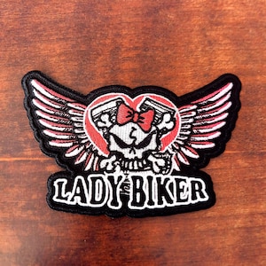 May include: Embroidered patch with a skull design, red bow, and wings. The patch features the words "LADY BIKER" in white on a black background. The wings are red and white, with a heart-shaped design.