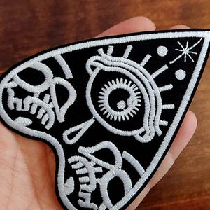 May include: Black and white embroidered patch in a heart shape. The patch features a stylized eye, skull, and geometric designs. The design is outlined in white thread against a black background.
