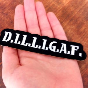 May include: Black embroidered patch with the text "D.I.L.L.I.G.A.F." in white. The patch is rectangular with rounded edges. The background is a wooden surface.