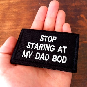 May include: Black rectangular patch with white embroidered text reading "STOP STARING AT MY DAD BOD". The patch is held in a hand.