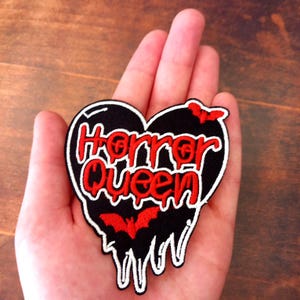May include: A black heart-shaped patch with the words "Horror Queen" in red, outlined in white. The patch features red bat silhouettes and a dripping design, ideal for adding to clothing or accessories.