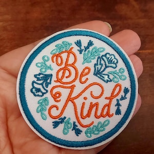 May include: Circular embroidered patch featuring the words "Be Kind" in orange script, encircled by teal and blue floral patterns. The patch has a white background and a teal border.