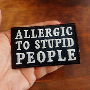 Puede incluir: Un parche rectangular negro con texto bordado en blanco que dice "ALLERGIC TO STUPID PEOPLE". El parche se sostiene en una mano sobre un fondo de madera. Un accesorio humorístico.